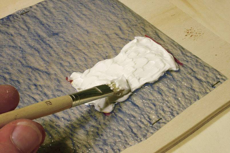 Un pinceau étale une pâte adhésive blanche sur une surface structurée bleu et beige. La main à gauche tient le pinceau.