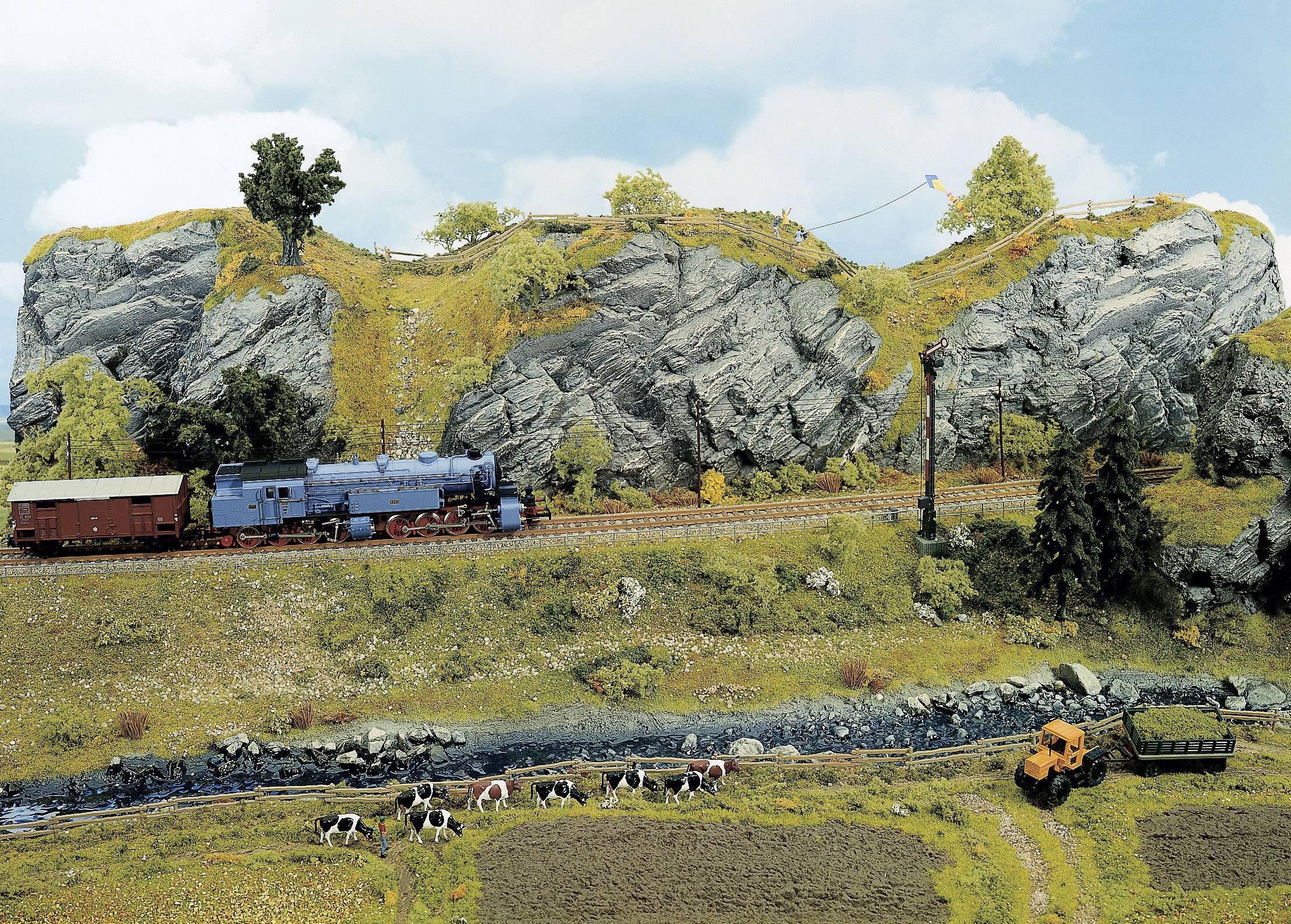 Un train miniature traverse un paysage rural avec des collines, des rochers et des prairies. Au premier plan, des vaches paissent.