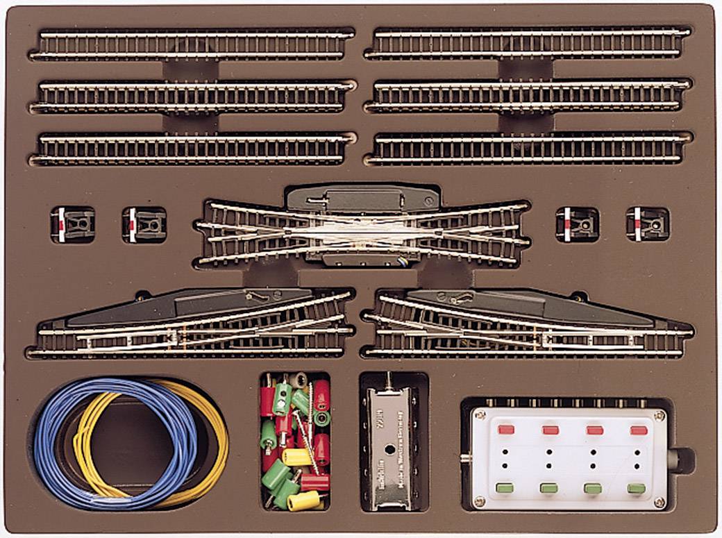 Ensemble d'accessoires pour train miniature comprenant des rails, des aiguillages, des câbles colorés, des interrupteurs et un panneau de contrôle, présenté dans son emballage ouvert.
