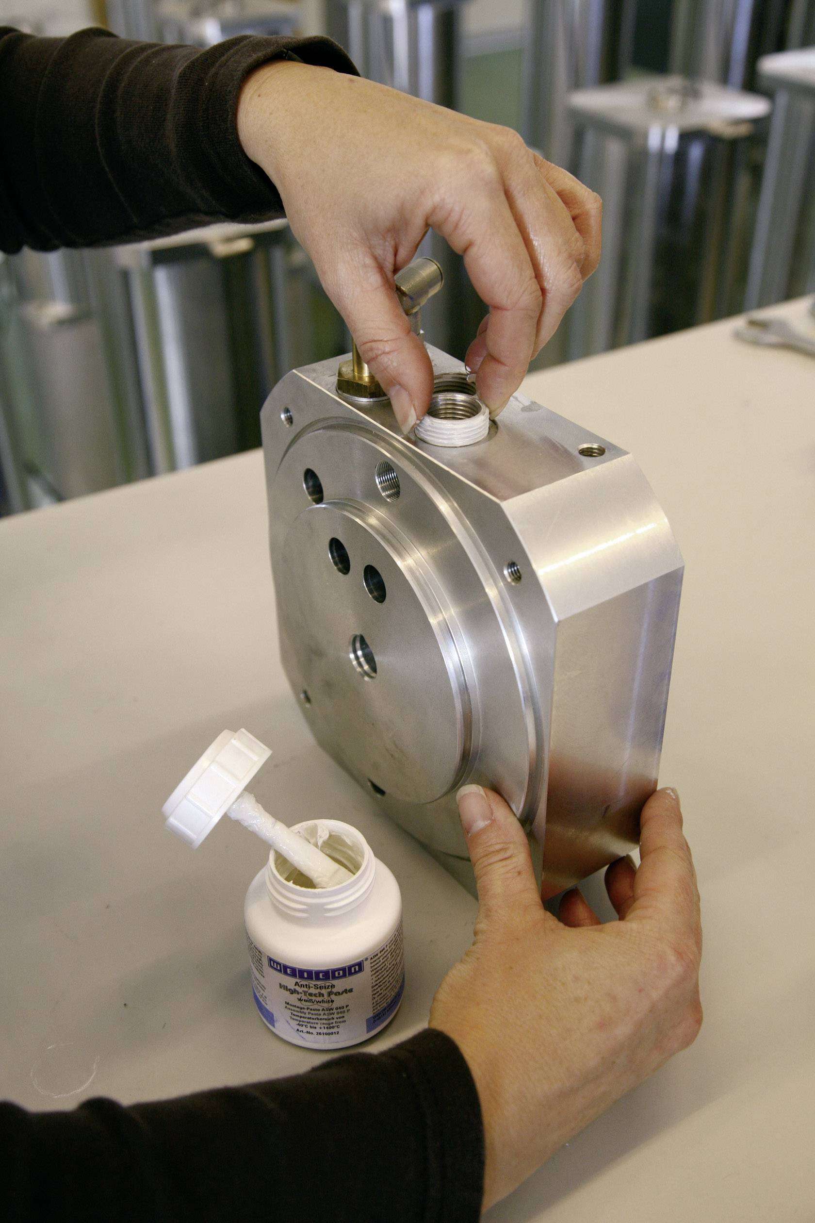 Une personne assemble une pièce métallique à l'aide de vis. Une bouteille ouverte de mastic est posée à côté sur l'établi.