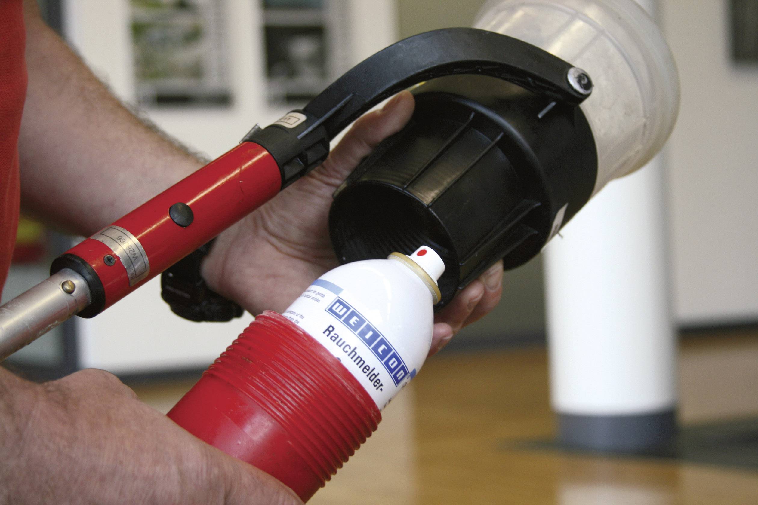 Une personne teste un détecteur de fumée à l'aide d'une bombe aérosol. La bombe est rouge et blanche et est dirigée contre le détecteur.