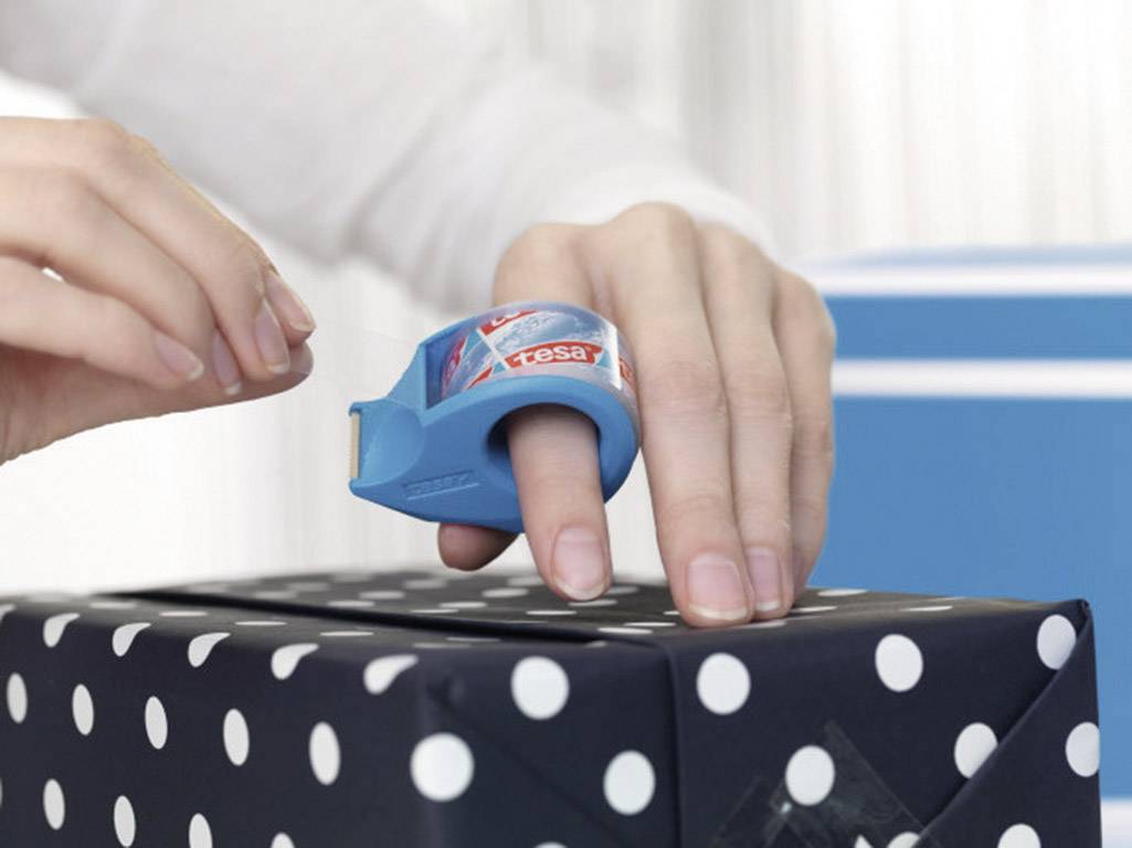 Une main utilise un distributeur de ruban adhésif bleu pour emballer un cadeau avec du papier d'emballage à pois.