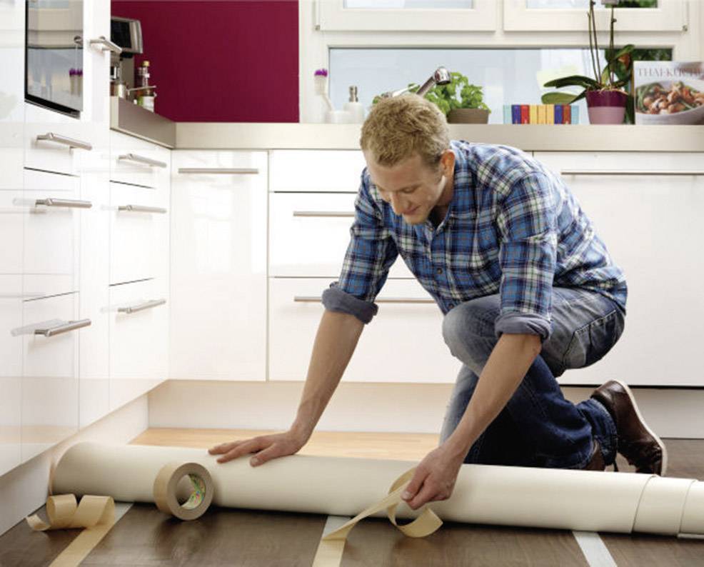 Un homme portant une chemise à carreaux installe un tapis de sol dans une cuisine moderne. Il déroule le matériau avec soin.