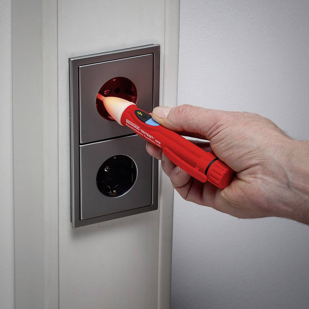 Une main tient un testeur de tension contre une prise électrique pour mesurer la tension. Le testeur affiche un voyant rouge comme signal d'avertissement.
