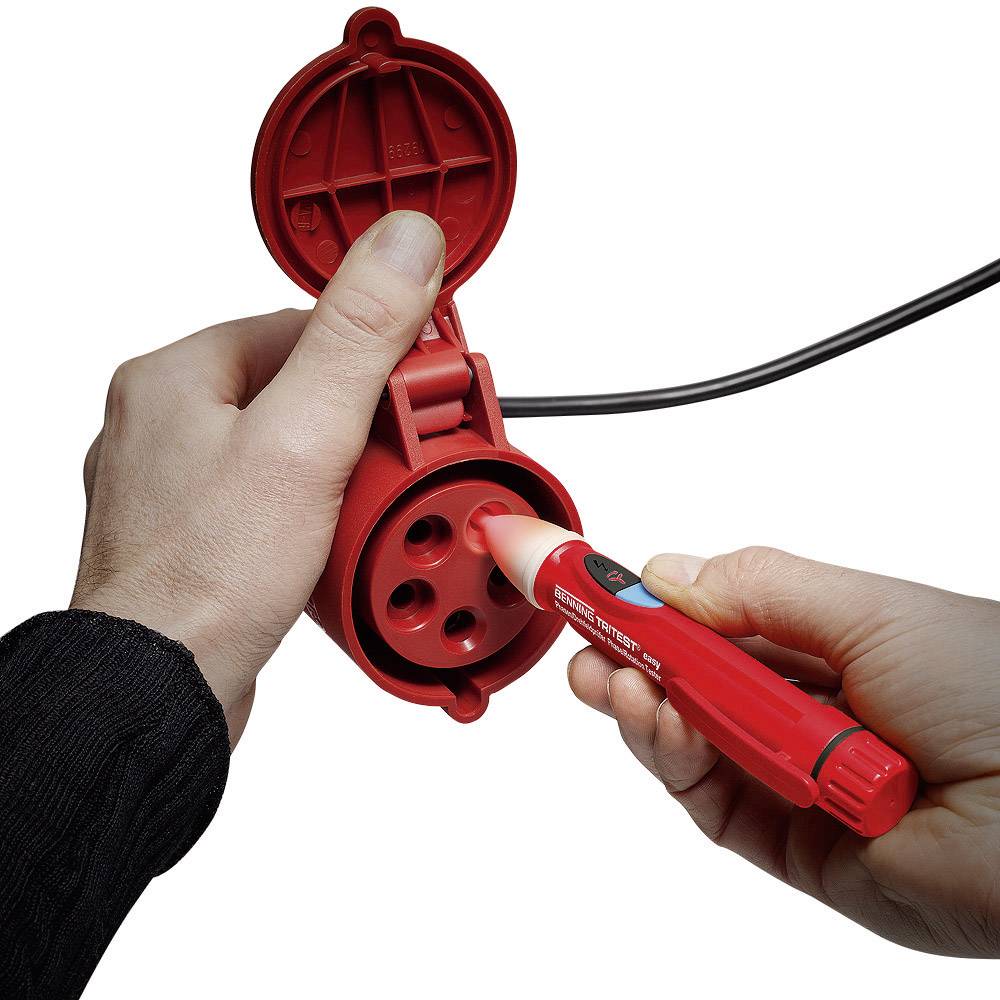 Une personne tient un testeur de tension près d'une prise électrique rouge, ronde et tripolaire afin de vérifier la tension.