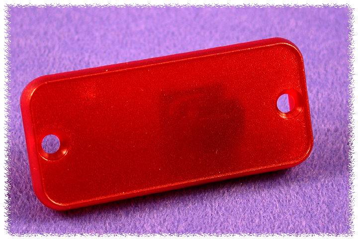 Une étiquette rectangulaire en plastique rouge avec des coins arrondis et deux trous, posée sur un fond lilas.