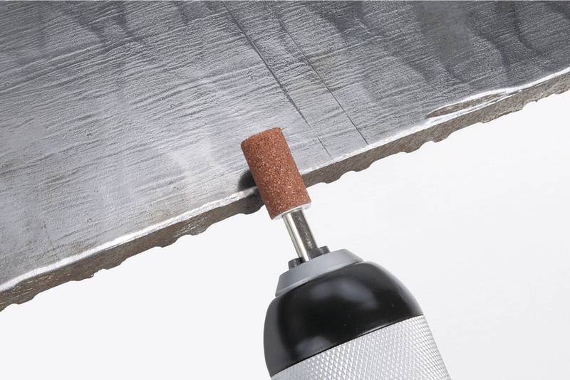 Un outil de meulage travaille le bord d'une plaque métallique. L'outil a une tête de meulage rouge-brune et polit la surface.