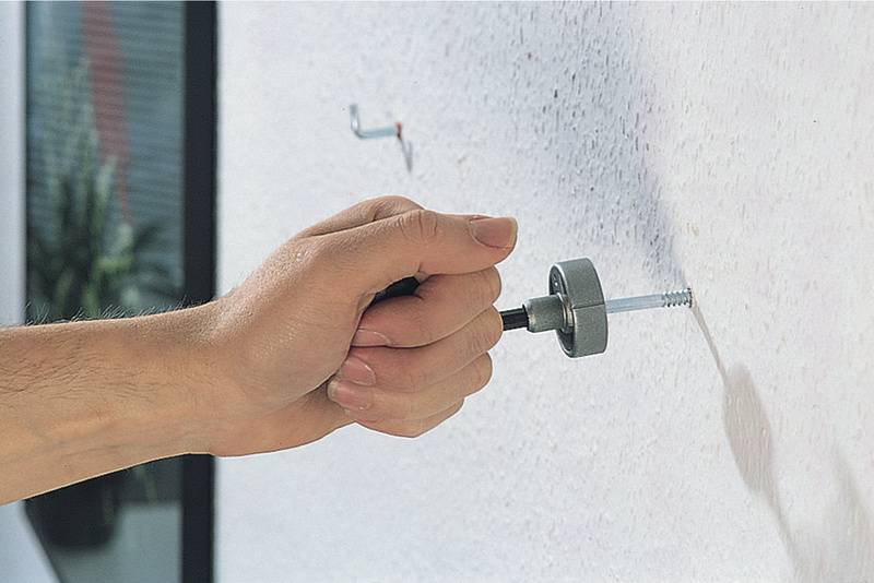 Une main tourne une grande vis dans un mur blanc à l'aide d'un outil. Une plante est visible en arrière-plan.
