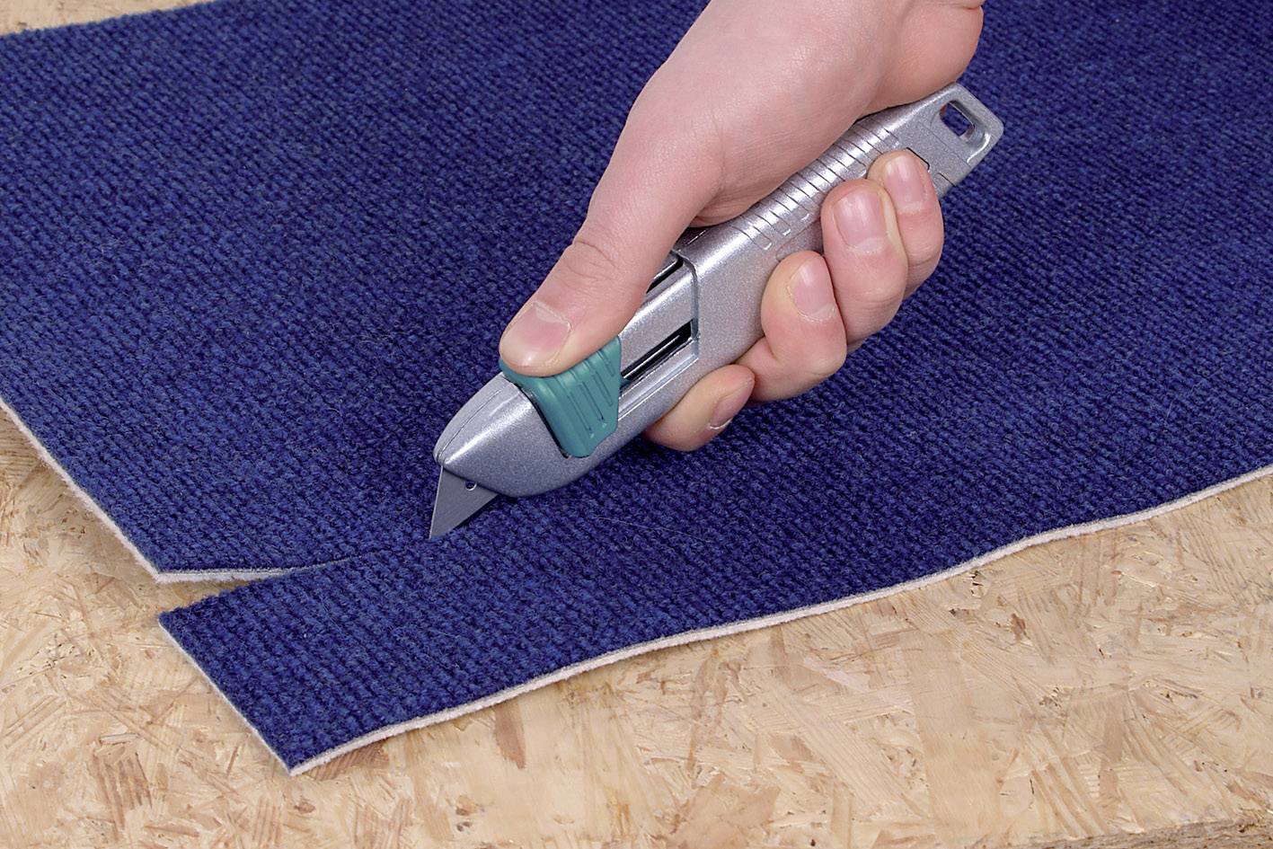 Une personne coupe un tapis bleu avec un couteau à tapis argenté sur une table en bois.