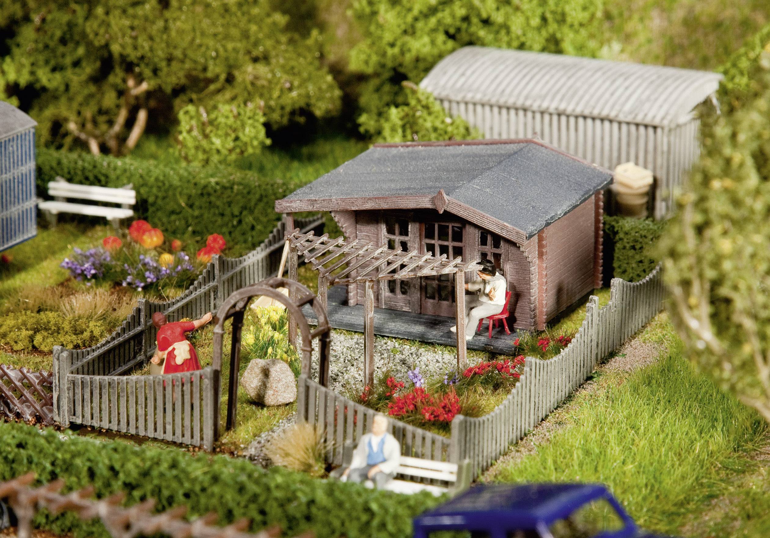 Un jardin miniature avec une petite maison, une clôture en bois et des bancs, entouré de fleurs et d'arbres en fleurs. Des figurines sont placées au premier plan.