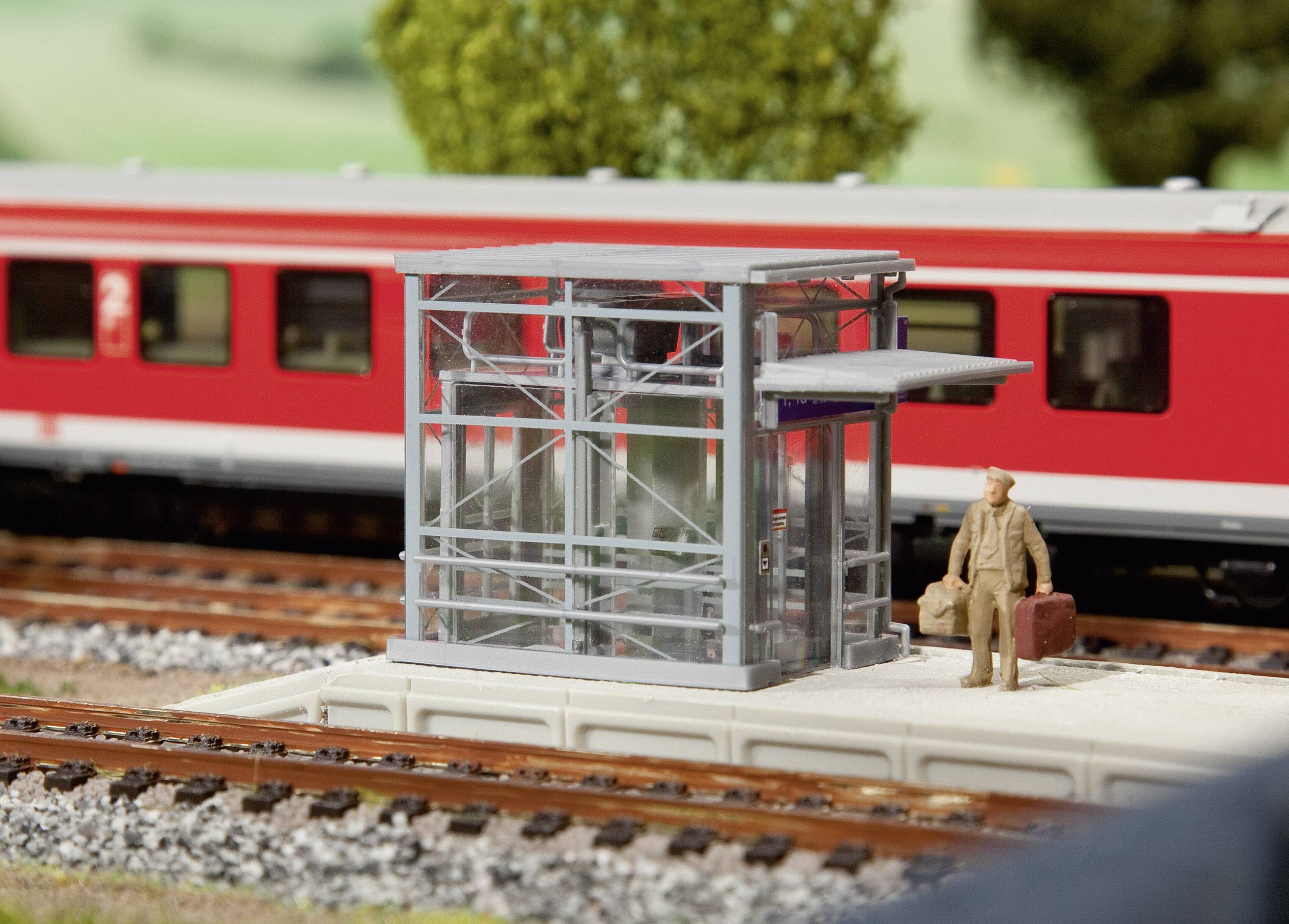 Une gare miniature avec un train rouge en arrière-plan. Au premier plan, une personne se tient avec des valises à côté d'un petit bâtiment en verre.