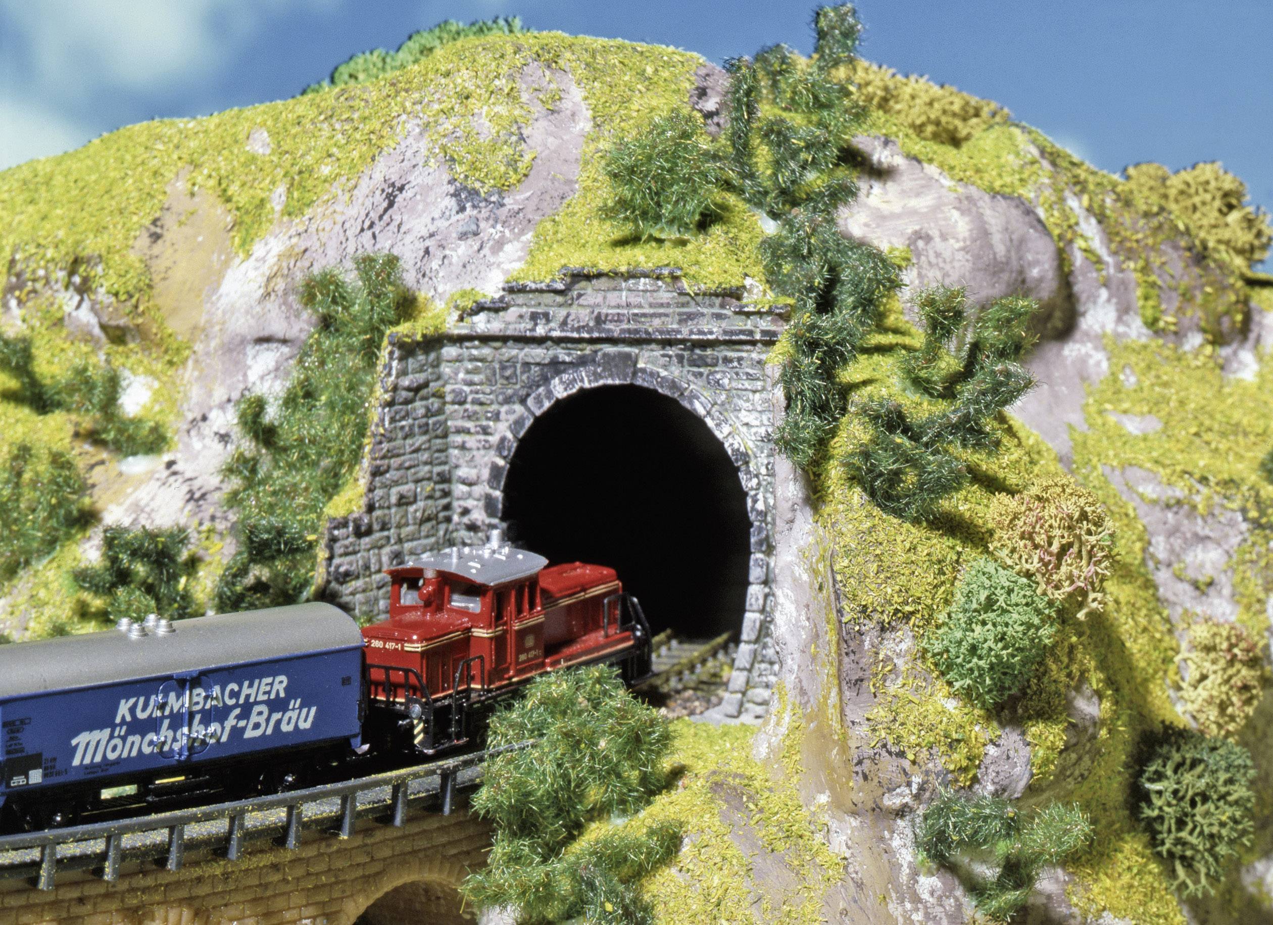 Un train miniature rouge et noir sort d'un tunnel dans un paysage miniature avec une colline verdoyante.