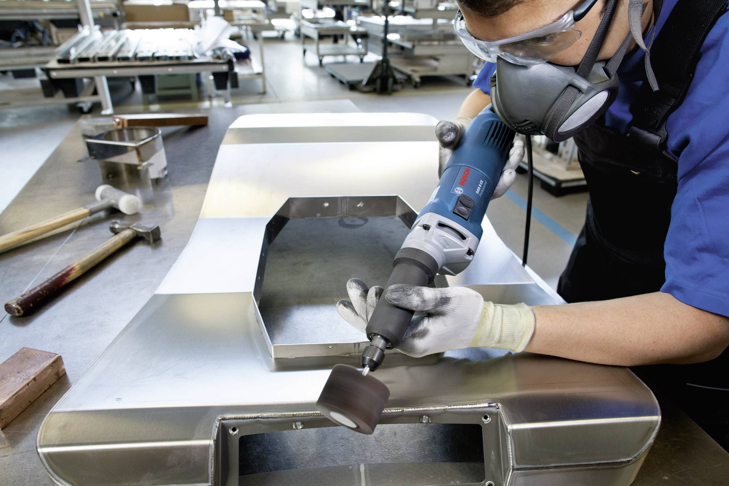 Une personne ponce une surface métallique avec une meuleuse électrique dans un atelier, portant des lunettes de protection et un masque respiratoire.