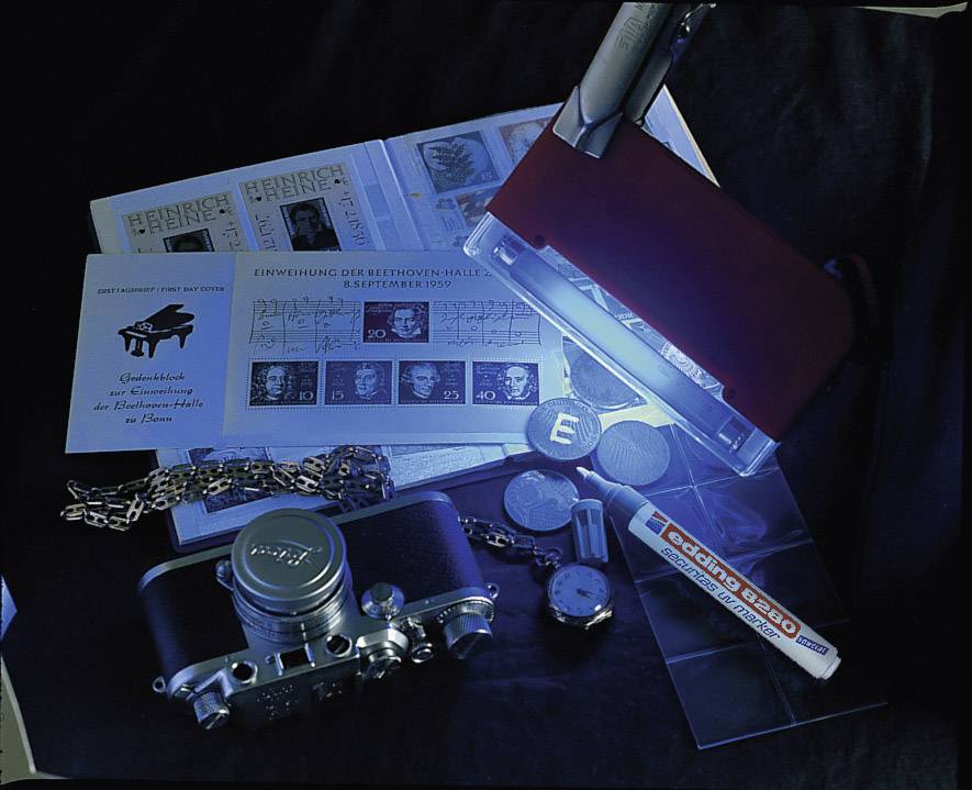 Un appareil photo, des montres, des bijoux, des timbres et une lampe UV sur une table. Des objets de collection et des outils de détection au premier plan.
