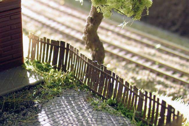 Paysage miniature avec une clôture en bois inclinée, un arbre et des rails de chemin de fer en arrière-plan ; illustrant une scène idyllique à petite échelle.