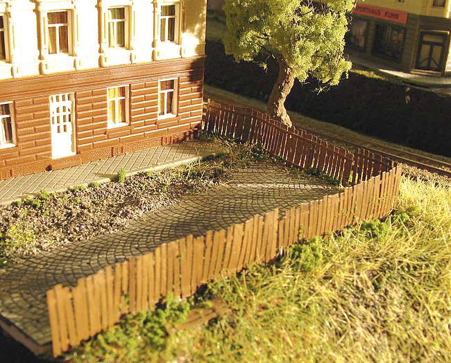 Scène de train miniature avec un bâtiment en briques, un vieil arbre et une clôture en bois sur un terrain herbeux.