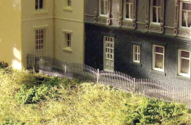 Plusieurs bâtiments miniatures aux façades détaillées sont alignés côte à côte. Une petite clôture ondulée sépare les bâtiments de l'herbe et des buissons en premier plan.