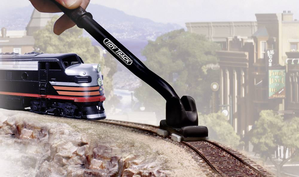 Une main nettoie une voie ferrée de maquette de train avec un outil, sur un paysage présentant des montagnes et des bâtiments en arrière-plan.