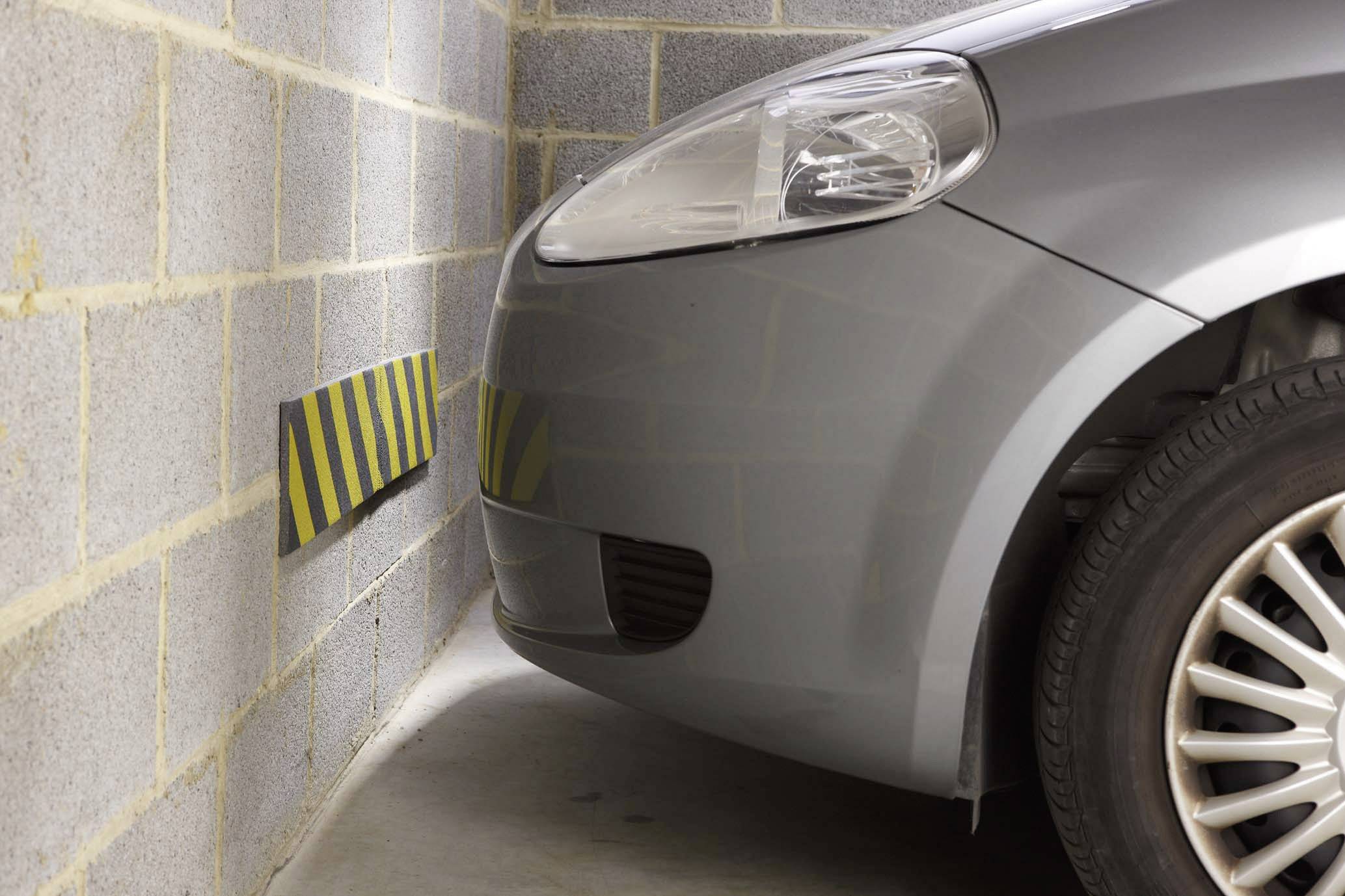 Une voiture grise est garée très près d'un mur dans un garage. Un panneau d'avertissement jaune et noir est accroché au mur.