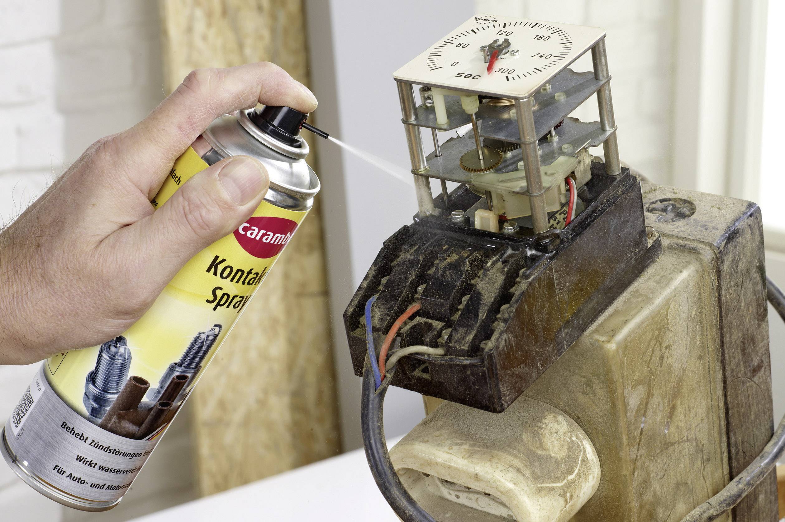 Une main pulvérise un spray de contact sur un composant électrique avec des fils afin de nettoyer les contacts électriques. L'objectif est d'améliorer la conductivité.