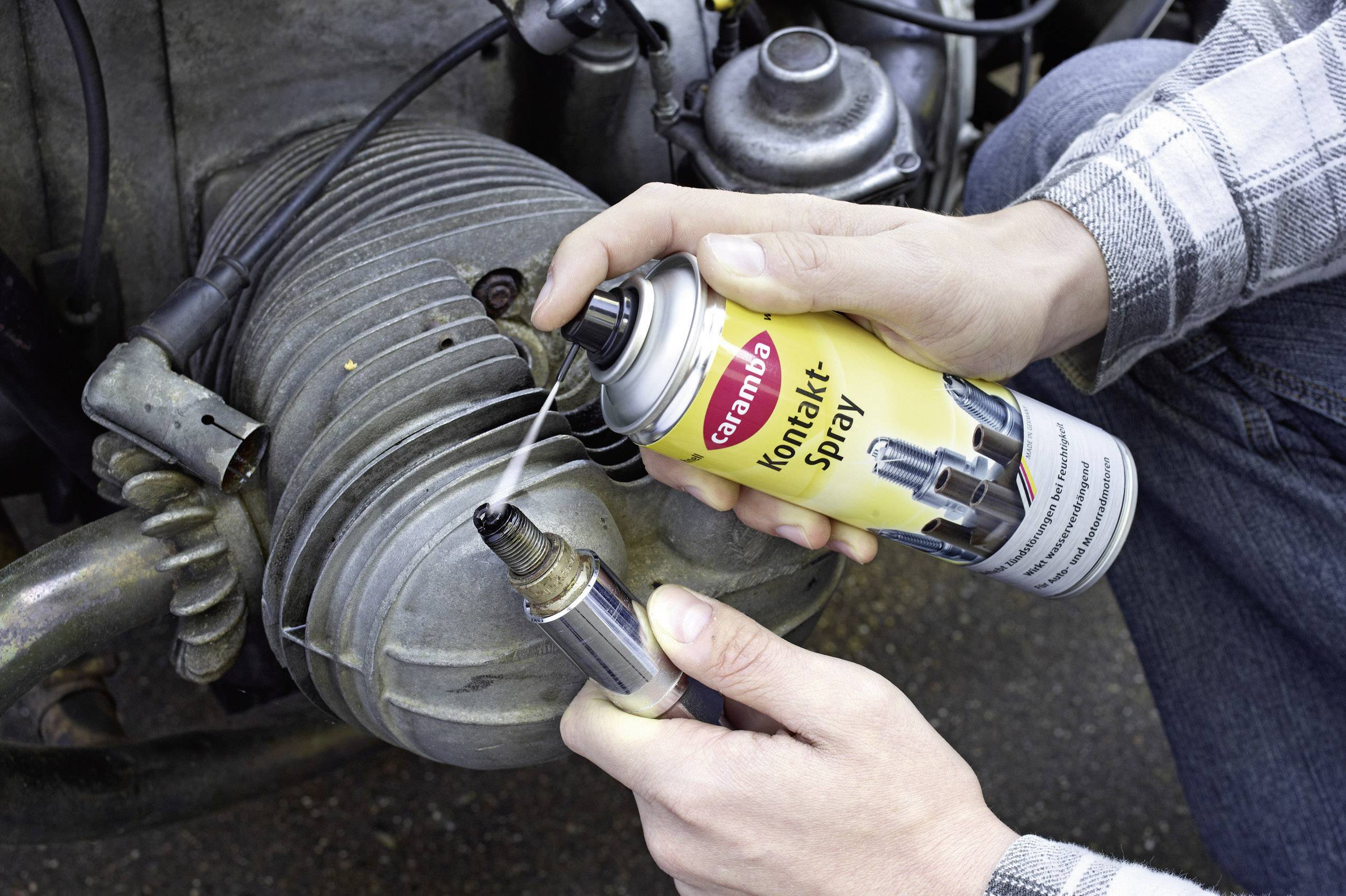 Une personne pulvérise un spray de contact sur une bougie d'allumage devant un bloc-moteur afin d'améliorer la conductivité électrique.