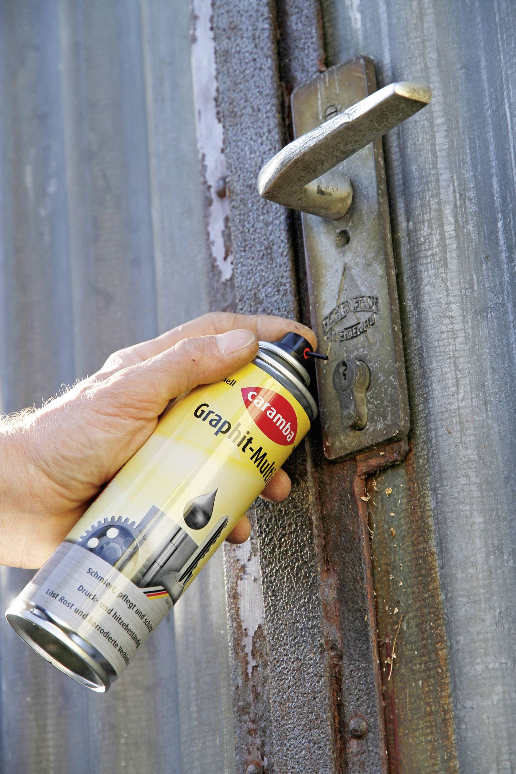Une main tient une bombe de 'Spray Huile Graphite' devant une charnière de porte rouillée, probablement pour la lubrification et l'entretien afin de réduire le frottement.