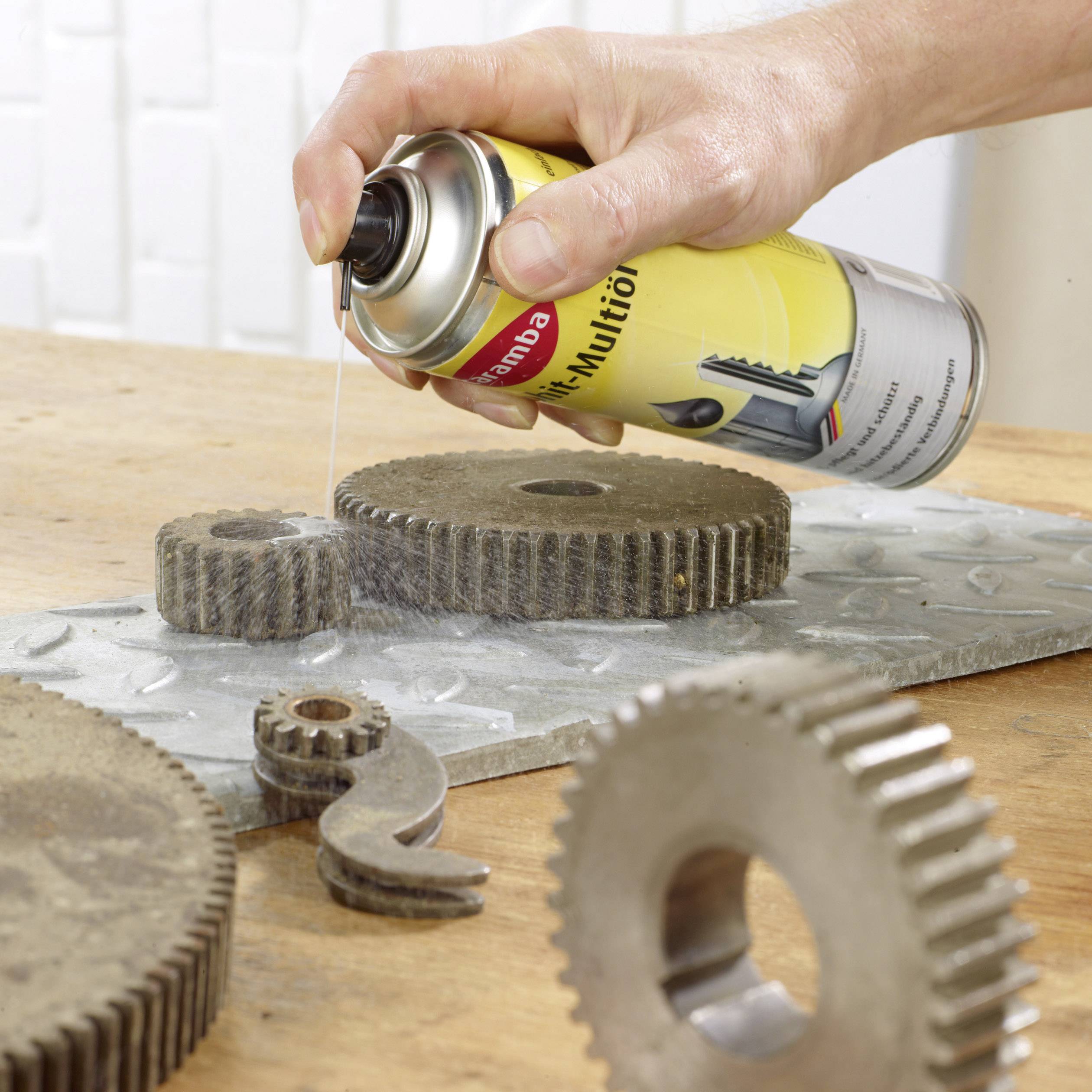 Une main pulvérise un lubrifiant sur des pièces de engrenage posées sur une table, afin de réduire la friction et d'améliorer la fonctionnalité.