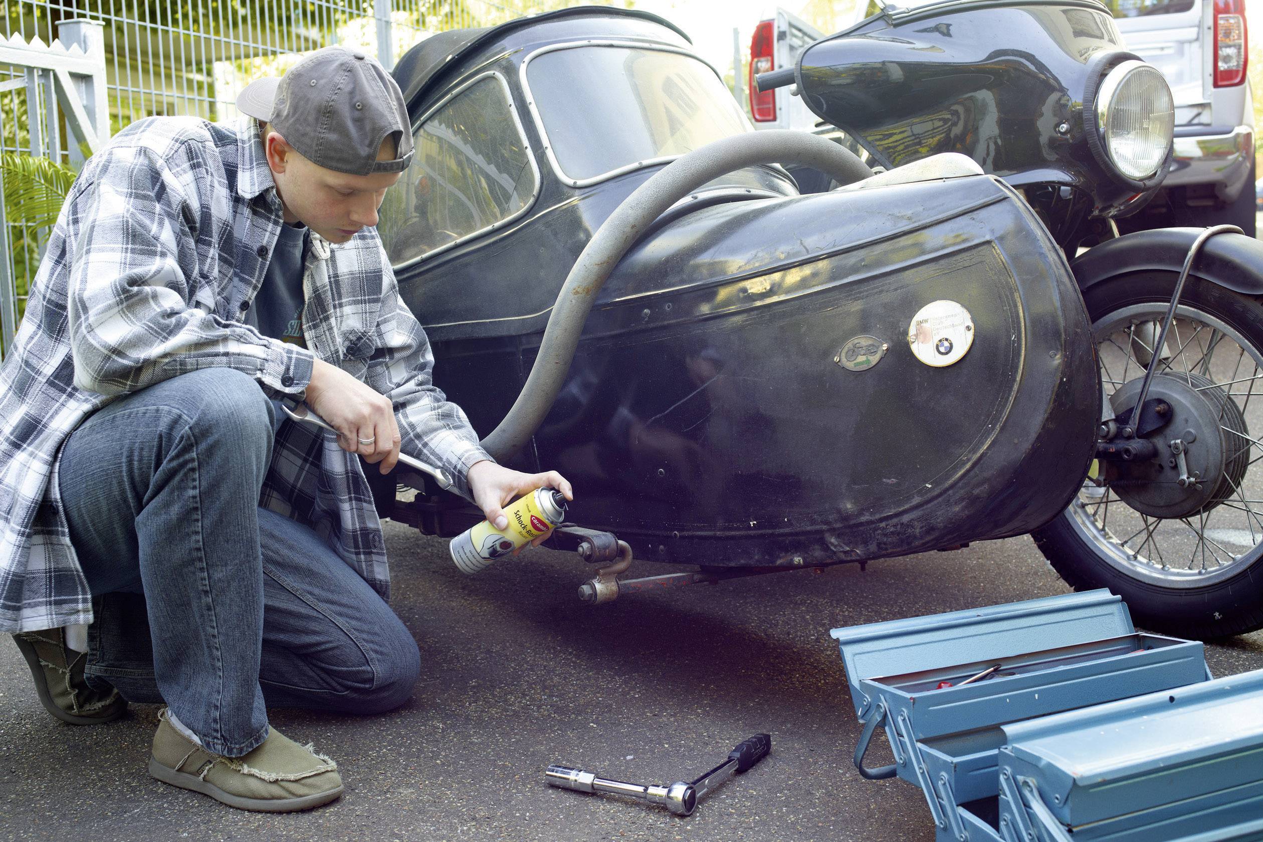 Un homme en chemise à carreaux et casquette à l'envers est à genoux à côté d'une moto avec side-car et pulvérise du lubrifiant sur des pièces du side-car.