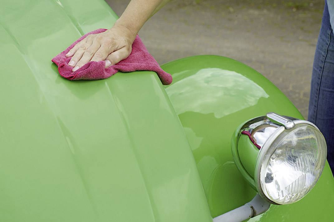 Une main polit le capot d'une voiture verte avec un chiffon rouge, mis en valeur par un détail de phare brillant.
