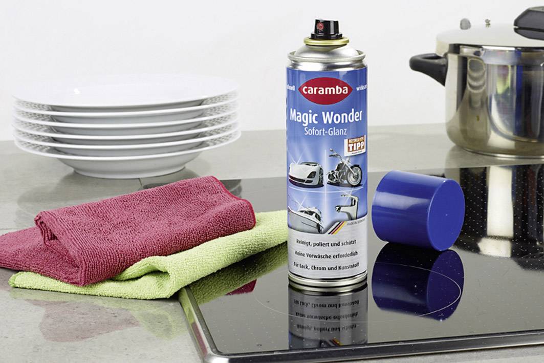 Un bidon de produit nettoyant 'Magic Wonder' est posé sur une cuisinière à côté de deux torchons de cuisine. En arrière-plan, des assiettes empilées et une casserole sont visibles.