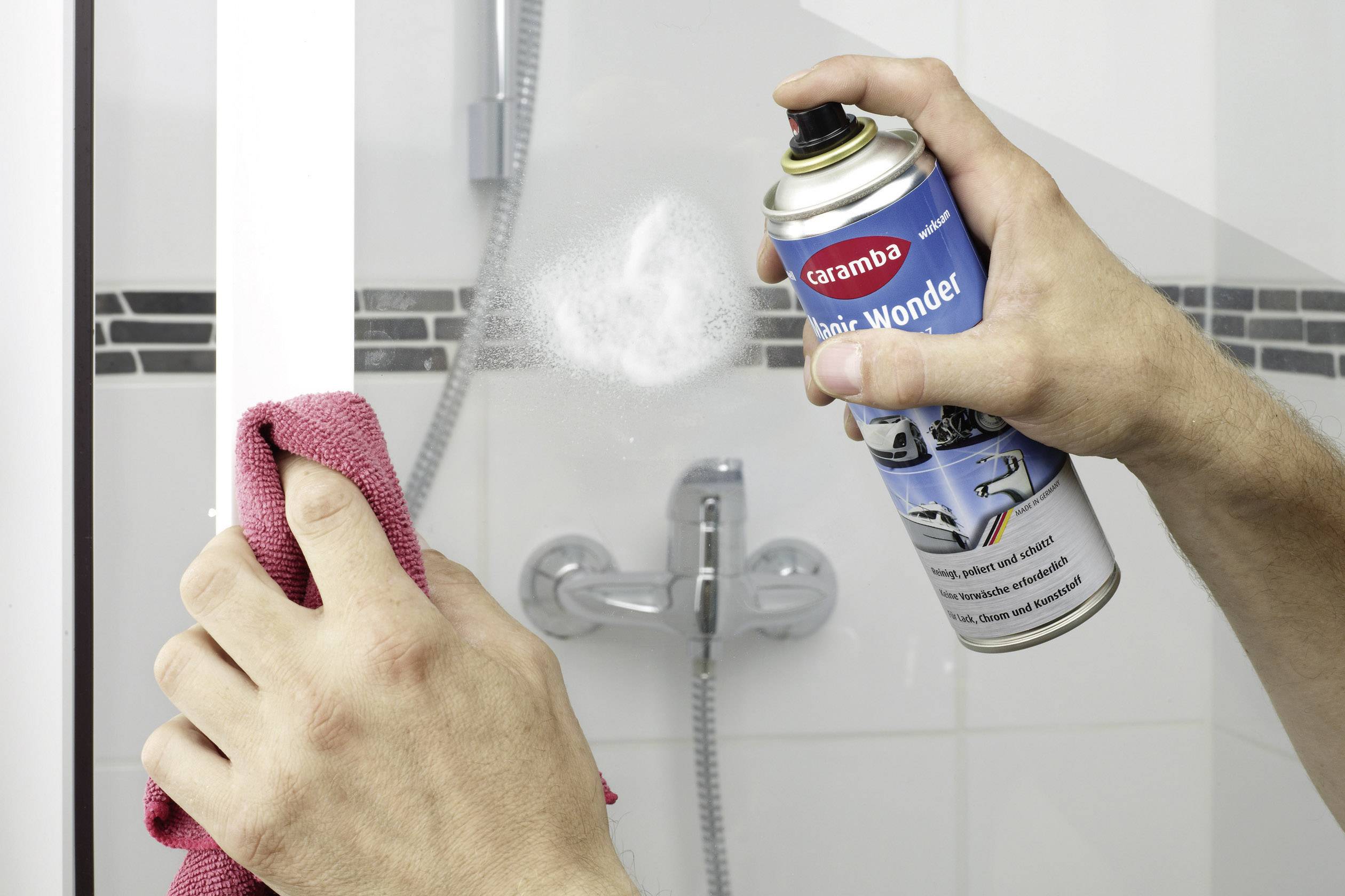 Une personne vaporise un produit de nettoyage sur une paroi de douche et essuie avec un chiffon rouge pour nettoyer la surface.