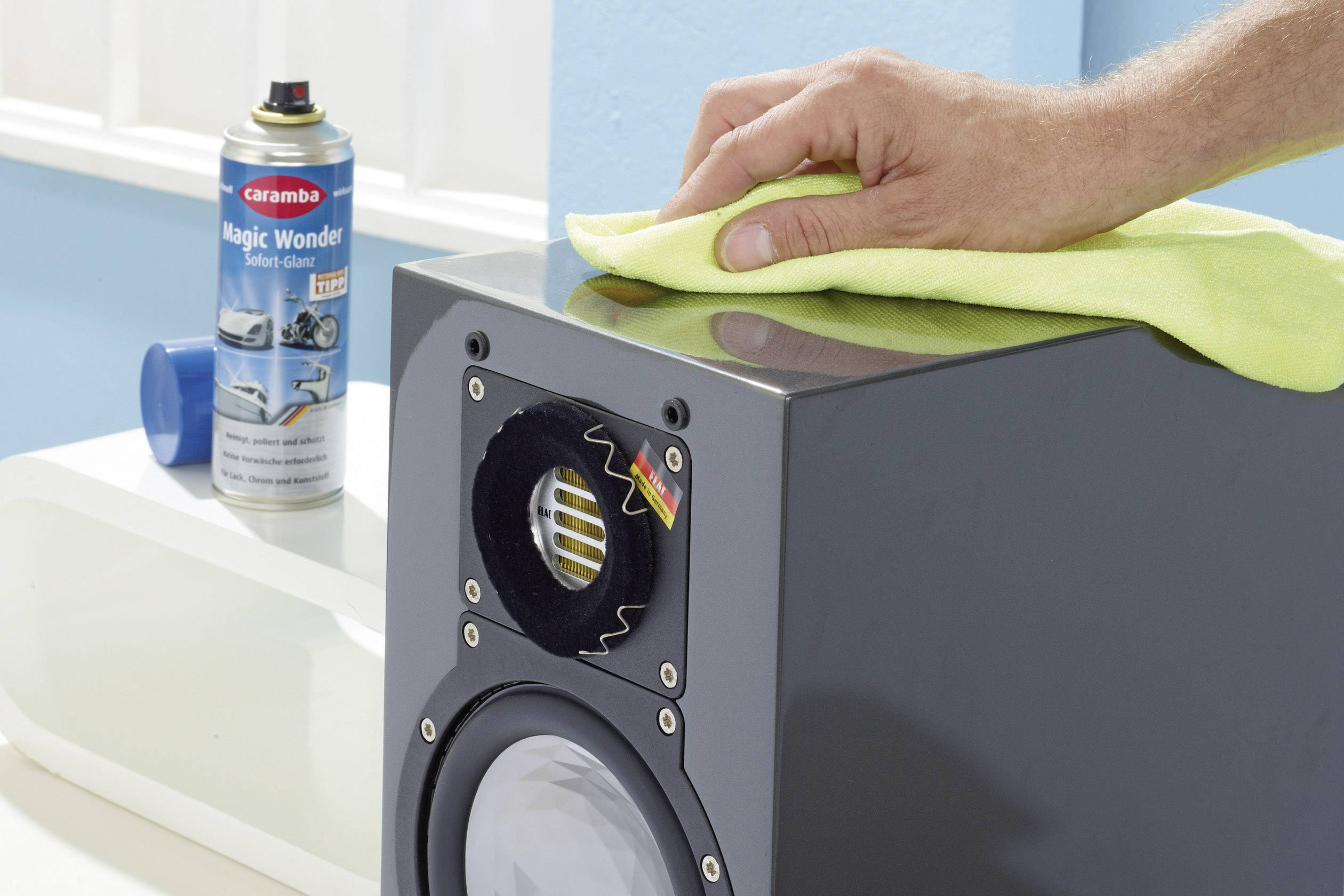 Une main essuie un haut-parleur avec un chiffon jaune. À l'arrière-plan, un aérosol de nettoyage pour voiture est posé sur une surface blanche.