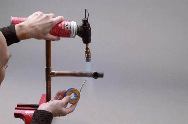 Une personne soude deux tubes de cuivre ensemble en tenant un chalumeau. Un tube est maintenu dans un étau.