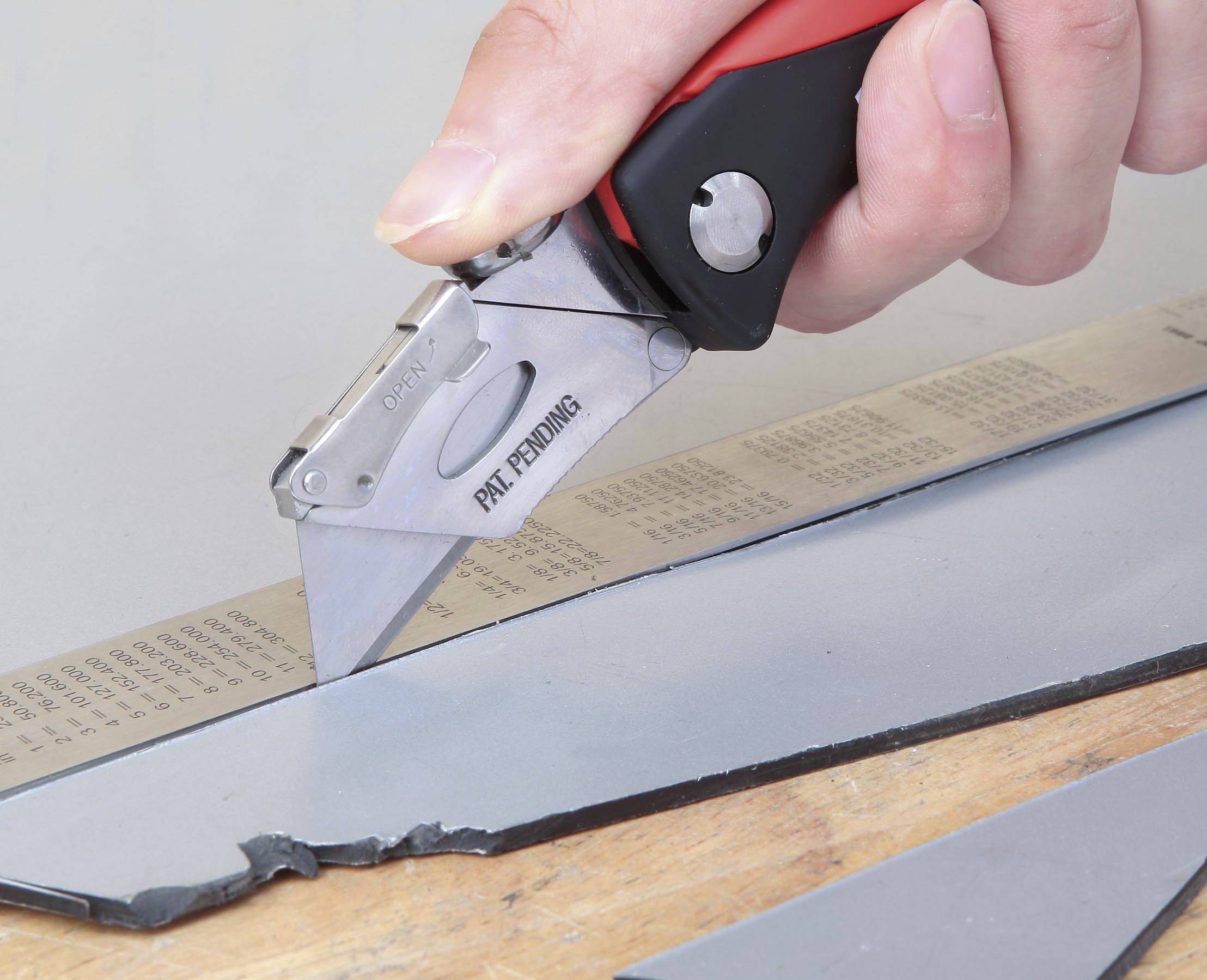 Une main coupe un morceau de matériau gris avec un cutter rouge sur une règle.