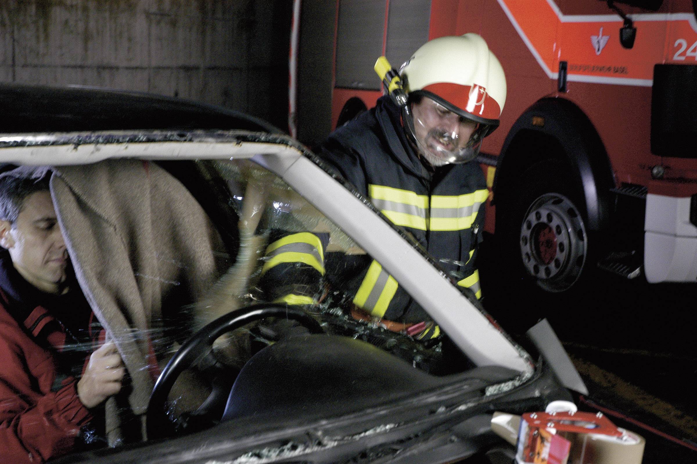 Un pompier sauve une personne d'une voiture accidentée. Un véhicule de pompiers est visible en arrière-plan.