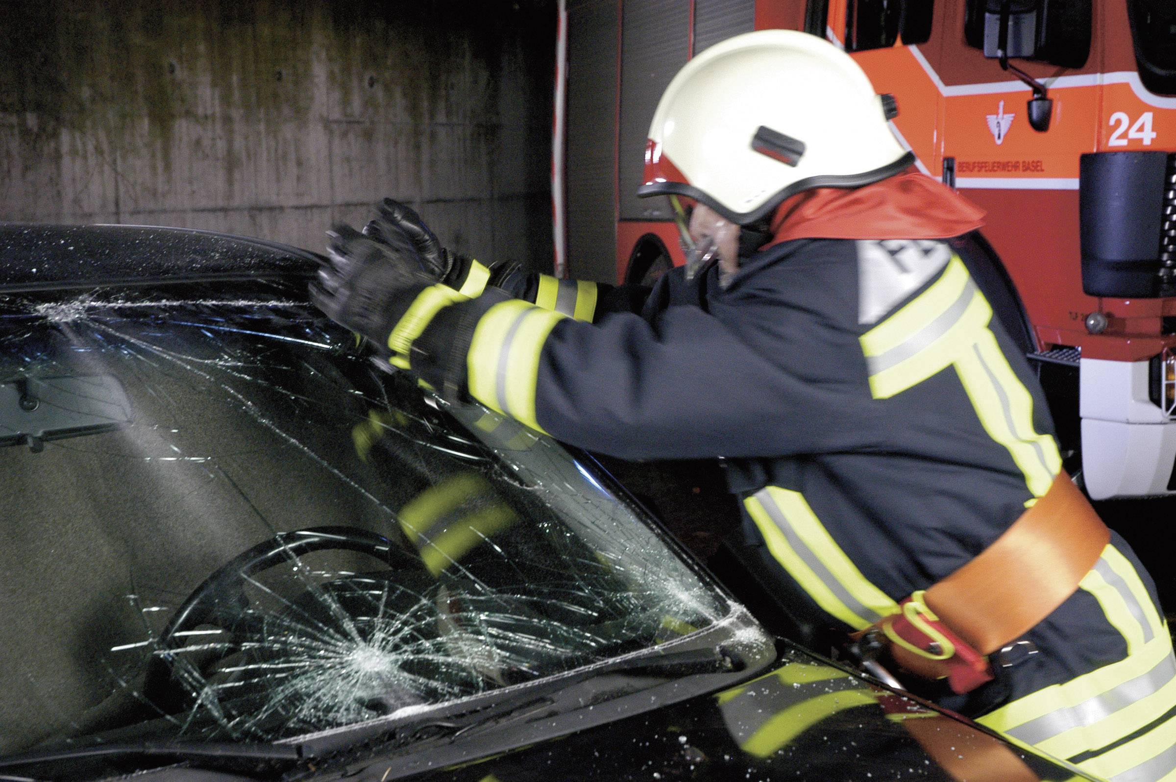 Un pompier en tenue de protection brise le pare-brise d'une voiture avec une hache. À l'arrière-plan, un camion de pompiers est stationné.