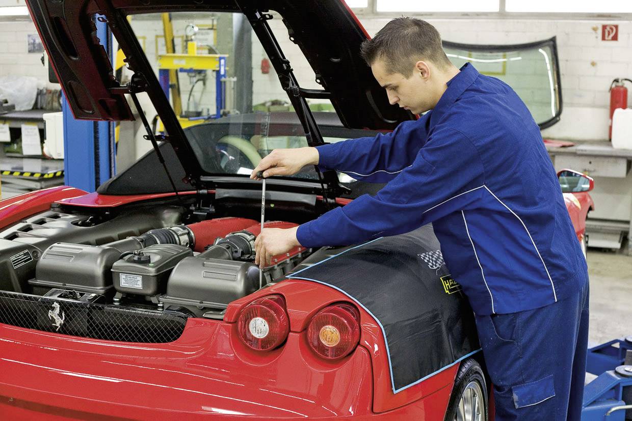 Un mécanicien en combinaison bleue travaille sur le moteur d'une voiture de sport rouge dans un atelier. Le coffre est ouvert.