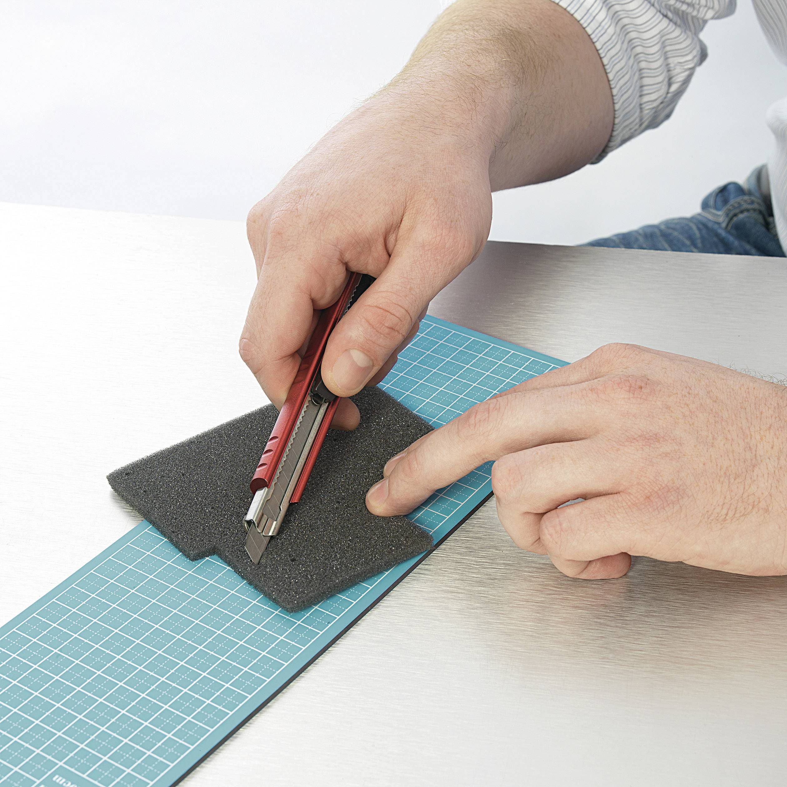 Une main découpe un morceau de mousse noire avec un cutter rouge sur un tapis de coupe.