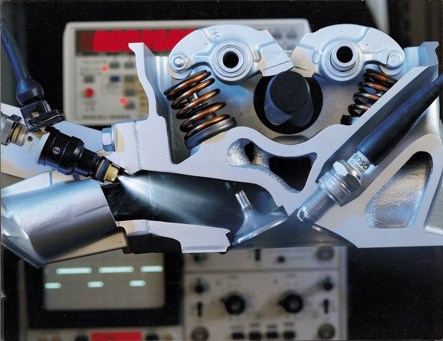 'Modèle en coupe d'un moteur automobile avec ressorts de soupapes et bougies d'allumage visibles. À l'arrière-plan, on peut voir des instruments de mesure avec des indicateurs lumineux rouges.'