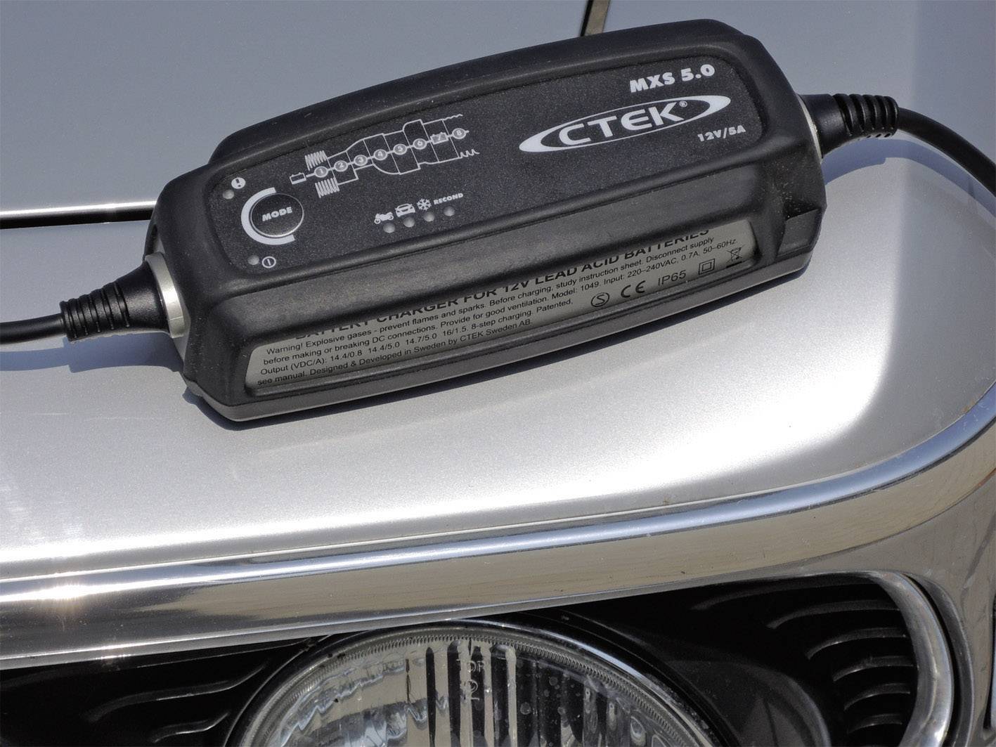 Le chargeur de batterie automobile 'CTEK MXS 5.0' est posé sur un capot de voiture. L'appareil affiche différents cycles de charge et voyants de fonctionnement.