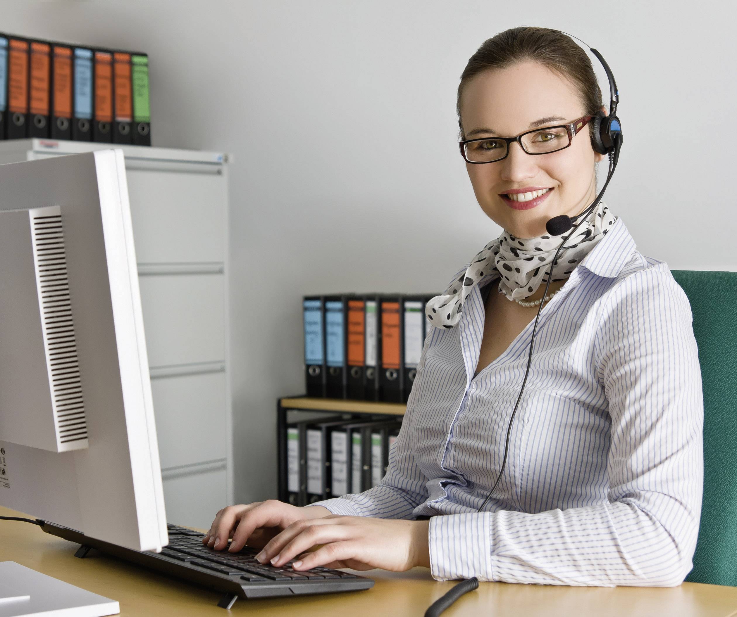 Une femme portant un casque est assise à un bureau devant un ordinateur. Elle sourit et porte une chemise rayée et un foulard. Des classeurs sont visibles en arrière-plan.